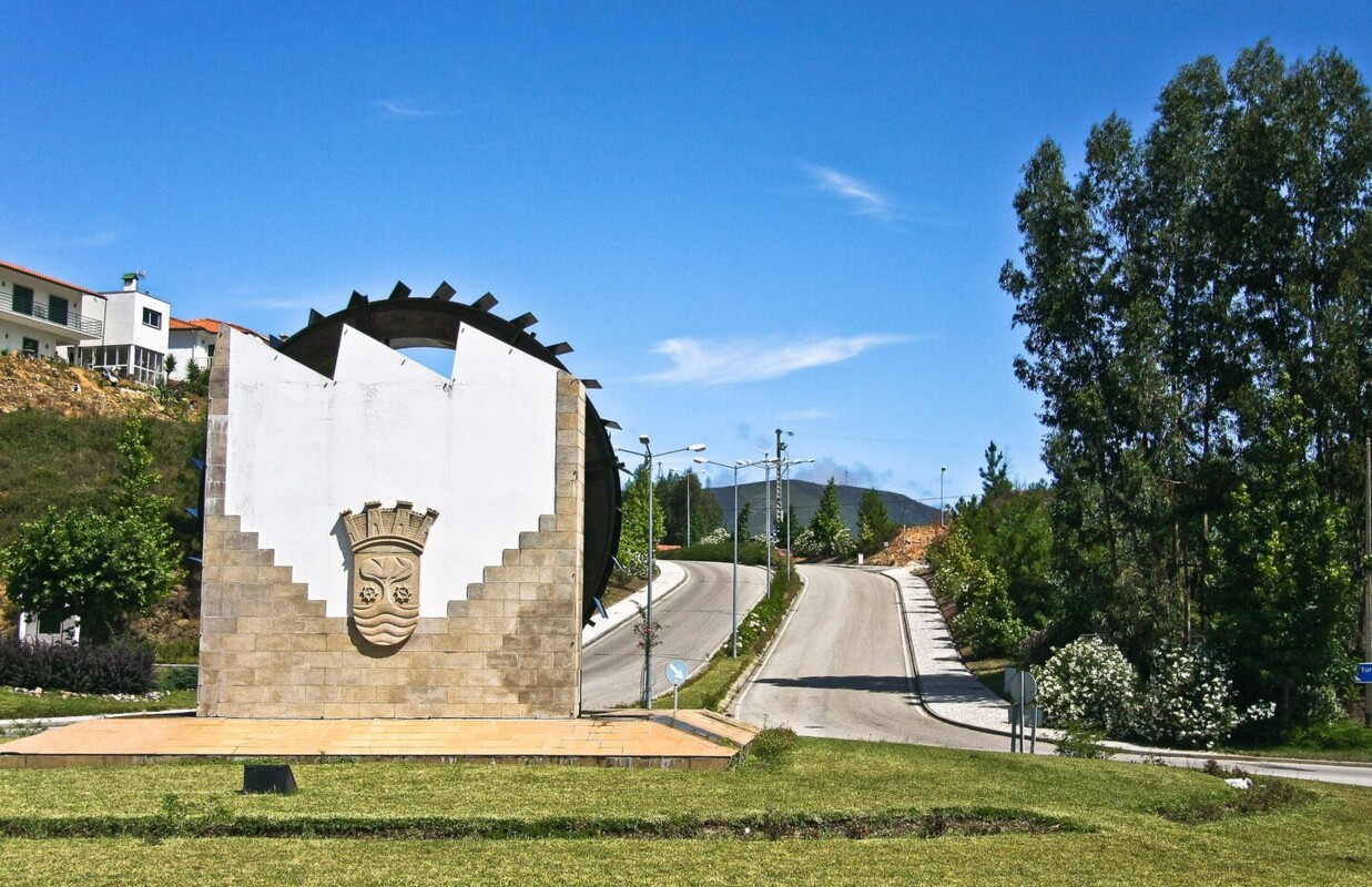 Brasão de Castanheira de Pera, numa parede em que a metade superior é branca e o restante é de pedra, num jardim. Atrás há duas estradas e árvores ao lado da estrada.