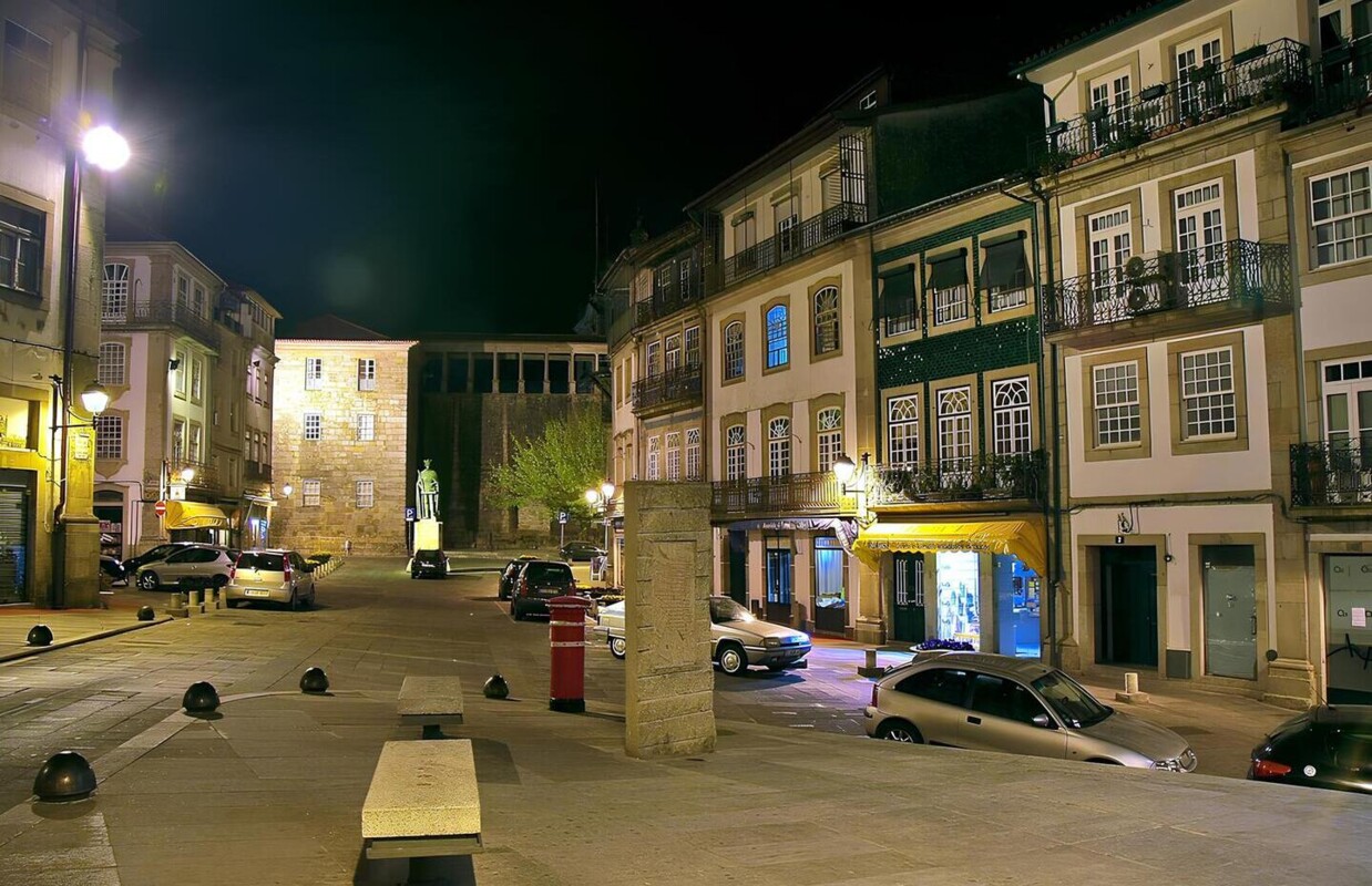 À noite, ao fundo, está uma estátua de ferro verde, numa avenida, com vários prédios antigos mas conservados de ambos os lados, um banco de pedra.