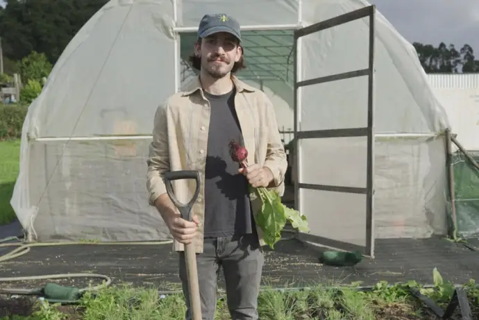 Tiago Fonteboa, fundador do projeto "Formiga Gloriosa" a segurar num engaço com a mão direita e com um rabanete na mão esquerda, com o terreno cultivado em pano de fundo