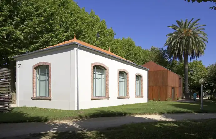 Edifício da nova biblioteca, branco com janelas, no meio de um jardim, ao lado de um edifício castanho.