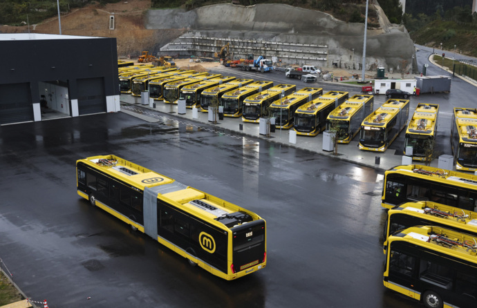 Vários autocarros amarelos, em fila virados para a frente, e um autocarro a dirigir-se em direção a outros três, que só se vê as traseiras, na diagonal, num espaço alcatroado, com um edifício preto.
