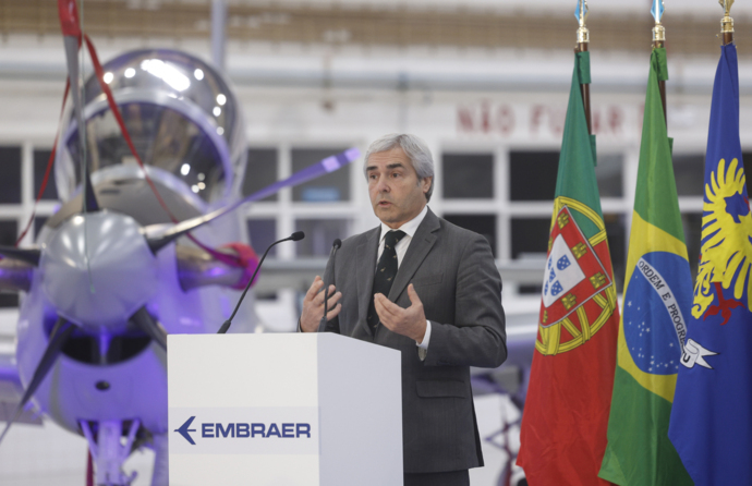 Nuno Melo, ministro da Defesa Nacional, ao centro, com uma aeronave Super Tucano atrás e a bandeira de Portugal e Brasil.