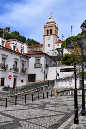 Uma rua da cidade de Leiria, onde estão alguns edifícios e a torre da igreja ao fundo, num dia limpo.