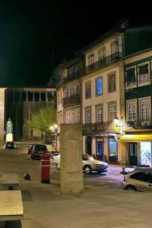 À noite, ao fundo, está uma estátua de ferro verde, numa avenida, com vários prédios antigos mas conservados de ambos os lados, um banco de pedra.