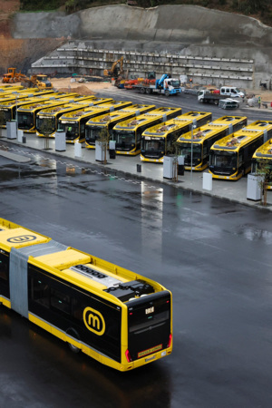 Vários autocarros amarelos, em fila virados para a frente, e um autocarro a dirigir-se em direção a outros três, que só se vê as traseiras, na diagonal, num espaço alcatroado, com um edifício preto.