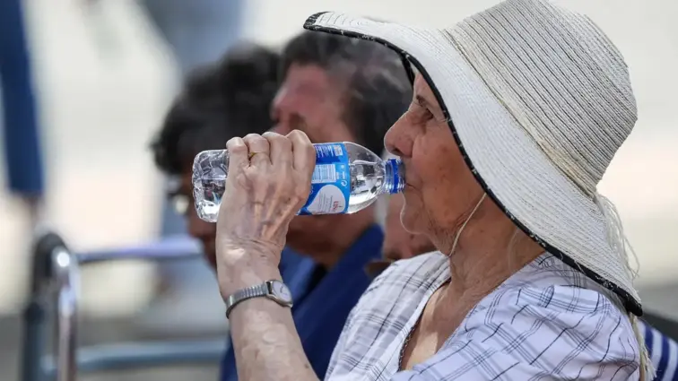 Mulher idosa a beber água e com chapéu na cabeça.