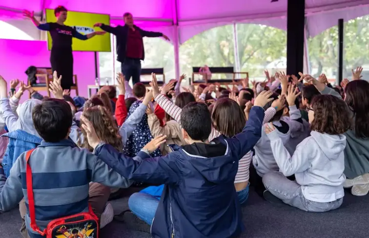 Crianças sentadas no chão, de costas, com as mãos no ar, a participarem de uma atividade do festival de literatura infantojuvenil de Valongo.