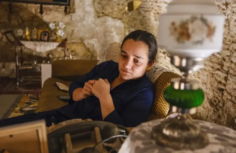 Uma atriz sentada num sofá, atrás de um candeeiro de mesa, na peça de teatro.