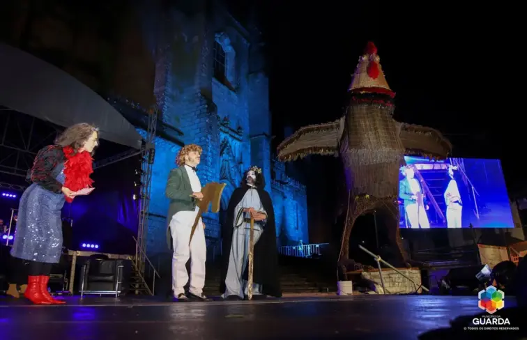 Pessoas e galo de madeira, tradicional do carnaval, num palco.