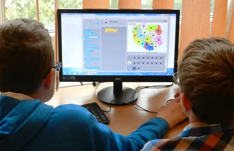 Duas crianças, de costas, a olhar para o monitor do computador onde estão a aprender programação.