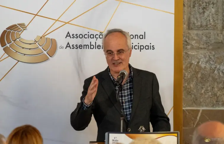 Albino Almeida, homem calvo de cabelo branco, vestido com um fato e camisa aos quadrados a discursar num palanque com microfone. Atrás, um placar com o logótipo de 'Associação Nacional de Assembleias Municipais'.