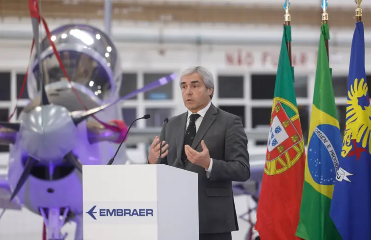 Nuno Melo, ministro da Defesa Nacional, ao centro, com uma aeronave Super Tucano atrás e a bandeira de Portugal e Brasil.