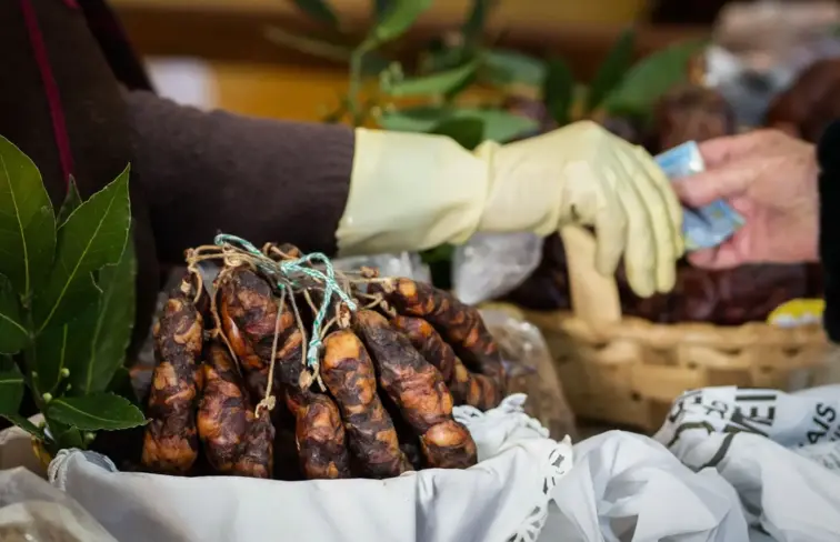 Imagem de enchidos expostos para venda, com detalhe de mão a receber dinheiro de uma venda