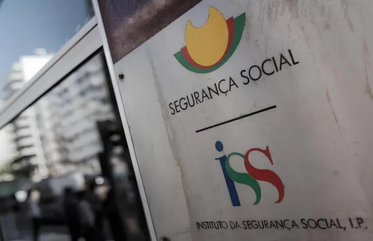 Plano aproximado de um placar num edifício, o placar é prateado e está escrito Segurança Social, com o respetivo logótipo em cima, e Instituto da Segurança Social, com o logótipo em cima, ao lado, há ainda o vidro de uma janela.