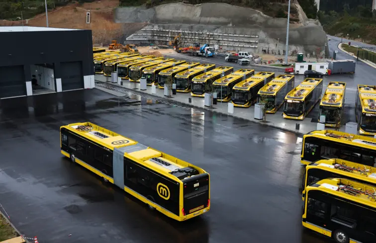 Vários autocarros amarelos, em fila virados para a frente, e um autocarro a dirigir-se em direção a outros três, que só se vê as traseiras, na diagonal, num espaço alcatroado, com um edifício preto.