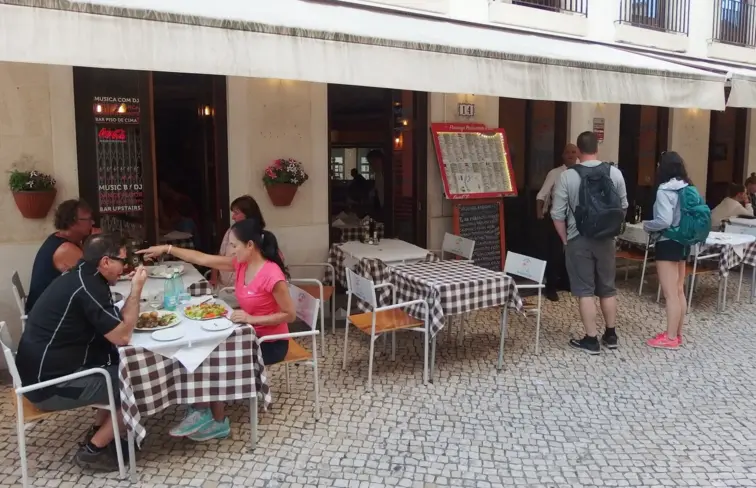 Uma esplanada de um restaurante, com mesas e pessoas a comerem e pessoas de pé.