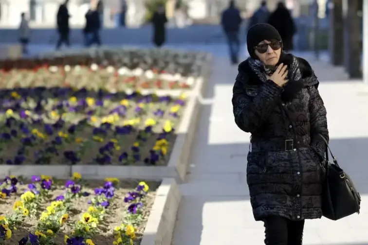 Mulher a caminhar na rua, junto a um jardim e muito agasalhada, a aconchegar o pescoço.