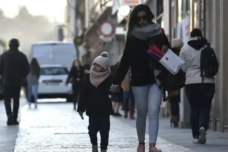 Uma mãe e um filho bem agasalhados de mãos dadas na rua, entre outros transeuntes agasalhados.