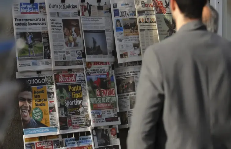 Homem a olhar para conjunto de jornais afixados num quiosque. 