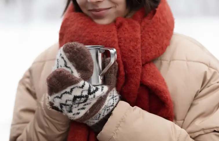 Mulher com casaco, cachecol e luvas, segura uma bebida quente no inverno.