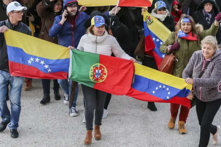 Três pessoas levam nas mãos três bandeiras: duas venezuelanas e, no meio, uma bandeira portuguesa. Atrás das três pessoas, uma multidão.