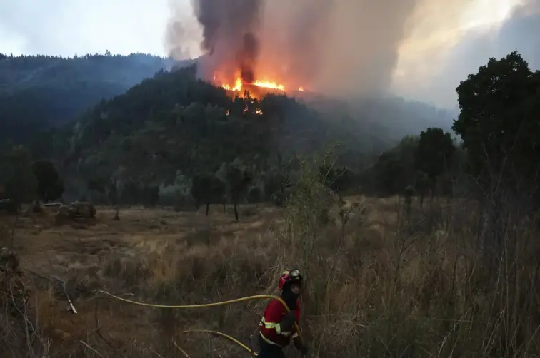 Serra em chamas ao fundo, num plano mais próximo vê-se um bombeiro.