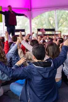 Crianças sentadas no chão, de costas, com as mãos no ar, a participarem de uma atividade do festival de literatura infantojuvenil de Valongo.