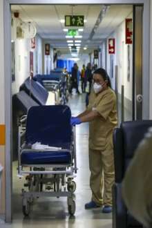 Corredor de hospital onde se vê uma profissional de saúde a transportar uma maca. 